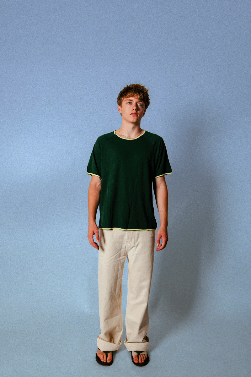 Model wearing the Soleil Lune Base Shirt in forest green with contrast stitching. Made from 55% hemp and 45% organic cotton, featuring a relaxed fit, lightweight natural texture, and soft drape styled with cream trousers in a minimal studio setting.