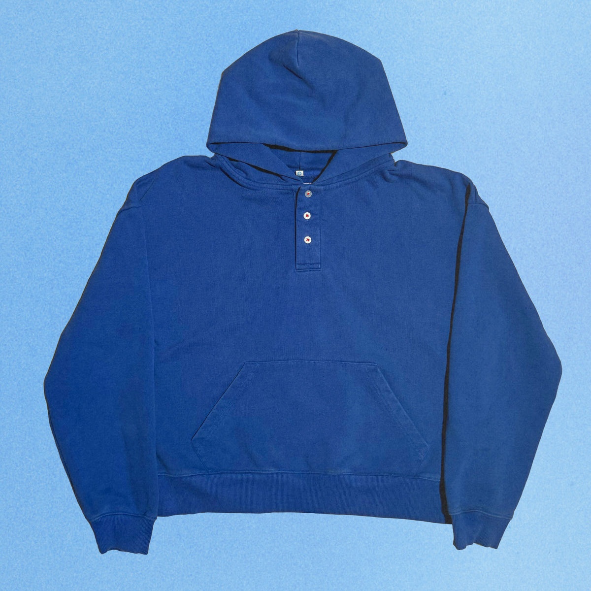 Blue heavyweight French terry Henley hoodie laid flat against a soft blue gradient background. The relaxed fit features a structured double layer hood, three button Henley placket with contrasting white buttons, ribbed cuffs and hem, and a spacious kangaroo pocket for an elevated everyday look.