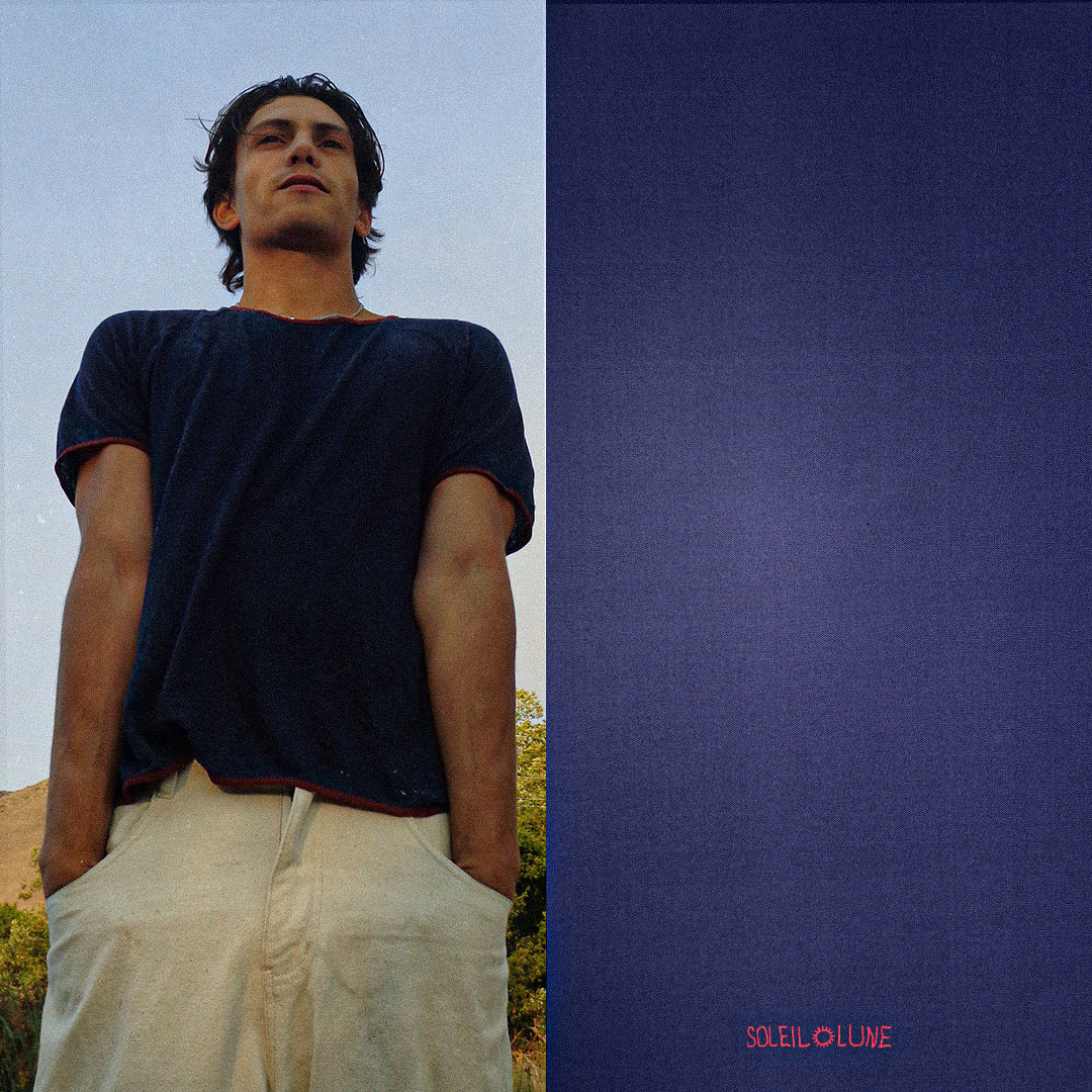 A young man stands confidently outdoors, wearing a navy blue t-shirt with red stitching and cream-colored pants. The background shows clear skies and trees, while the image is split vertically—on the right side, a textured deep blue background features small red text reading “SOLEIL LUNE” at the bottom center.
