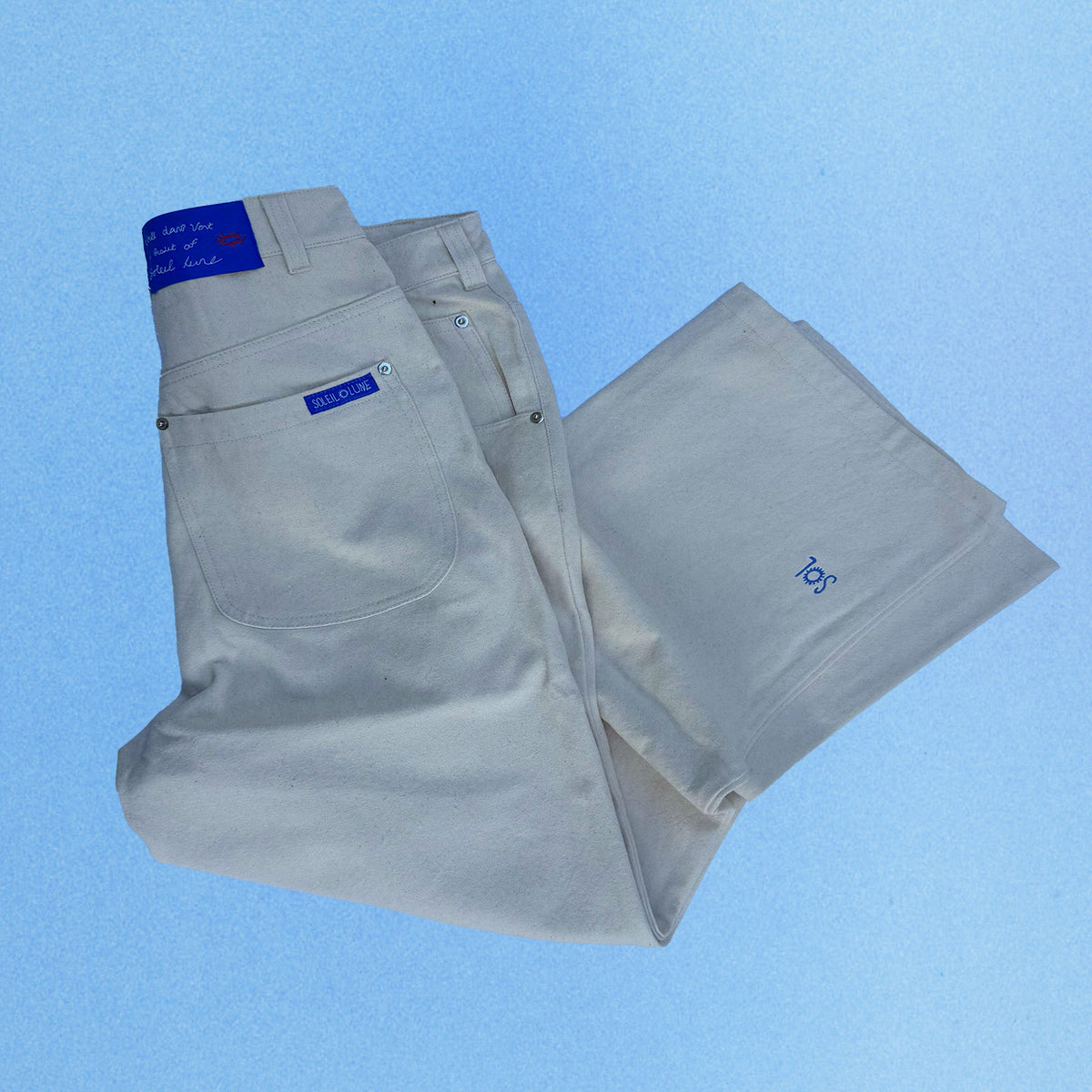 White heavyweight canvas pants folded flat against a soft blue gradient background. The back pocket features a blue woven Soleil Lune label, with additional blue branding on the waistband patch and subtle embroidered logo near the leg opening. Silver metal rivets and clean utility-inspired construction highlight the relaxed fit and durable craftsmanship.